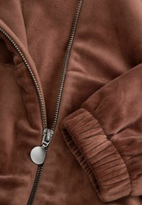 Zbliżenie na brązową miękką kurtkę polarową z metalowym zamkiem błyskawicznym i elastycznym mankietem na rękawie.