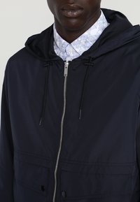 Navy hooded jacket with a smooth outer texture, silver zipper, adjustable drawstrings, and front pockets. Layered over a patterned shirt.