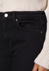 Svarta denimjeans med klassisk midja, med glänsande silverdetaljer, en framficka och en slät textur. Ribbad beige tröjärm synlig.