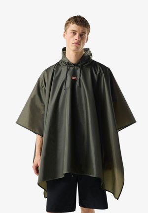 Mladić u tamnozelenom vodootpornom kišnom ponču s kapuljačom i vezicama, stoji ispred jednobojne bijele pozadine.