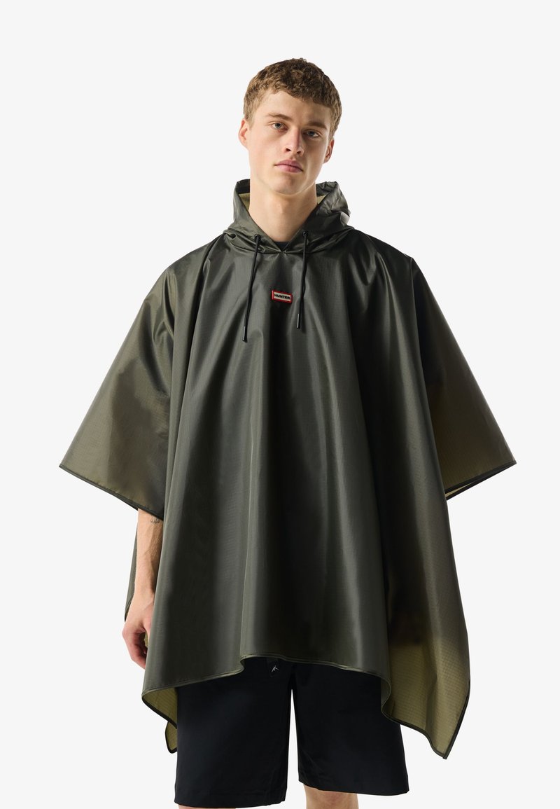 Νεαρός άνδρας που φοράει αδιάβροχο πονοφόρου σε σκούρο πράσινο χρώμα με κουκούλα και κορδόνια, στέκεται μπροστά σε λευκό απλό φόντο.