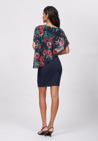Blommig genomskinlig kappa i marinblått med röda och rosa rosor, lager över en figursydd marinblå klänning. Modellen bär svarta sandaler med öppen tå.