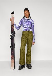 Suusatamisriided, mis sisaldavad pika varrukaga lilla ja valge mustriga särk, oliivrohelised veekindlad püksid, mustad suusasaapad ja lumelau.