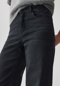 Jeans in denim nero con vestibilità slim, design a cinque tasche e dettagli di cucitura discreti, abbinati a un maglione in maglia grigio chiaro.