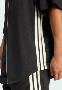 Čierna športová košeľa s priedušnou sieťovinou, krátkymi rukávmi a bielymi vertikálnymi pruhmi po stranách. Gombíky a kontrastné detaily lemovania.