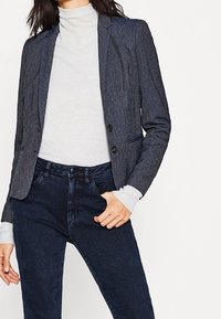 Námořnický blazer s texturovanou tkaninou, se zapínáním na jeden knoflík a dvěma předními kapsami, spárovaný se světle šedým rolákem a tmavými džíny.