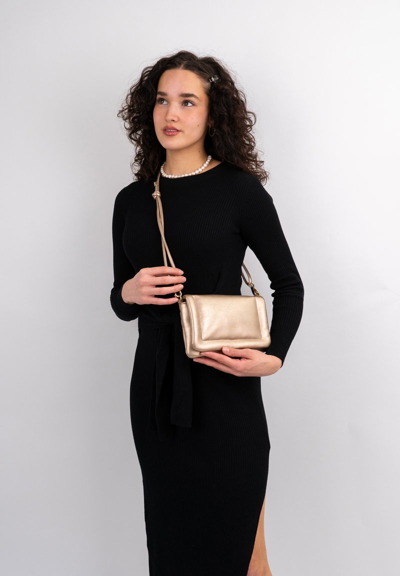 Borsa crossbody metallica dorata con una texture liscia, forma rettangolare e una tracolla staccabile, tenuta da una persona in un vestito nero.