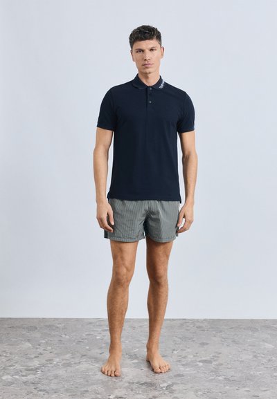 Tamsiai mėlynas polo marškinėliai su apykakle, puoštas šviesios spalvos siuvinėta detalė. Derinamas su pilkais ir baltais dryžuotais šortais. Neavint batų.