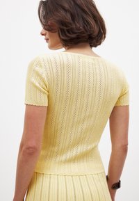 Geltona megzta trumpomis rankovėmis maikutė su vertikaliomis juostelėmis ir atviru raštu, turinti bangų formos kraštus prie rankovių ir apatinės dalies.