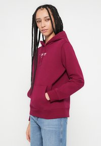 Μπορντό φούτερ με κουκούλα και τσέπη μπροστά, κορδόνι στην κουκούλα και ριμπ μανσέτες, φορεμένο με ανοιχτόχρωμο μπλε τζιν, διαθέτει λευκό λογότυπο κειμένου στο στήθος.