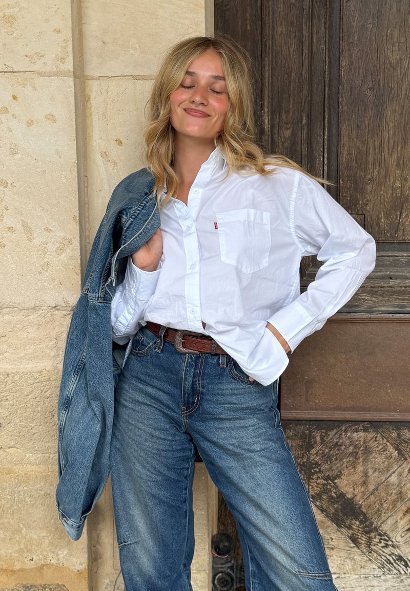 White long-sleeve shirt with a pocket, paired with high-waisted blue denim jeans and a denim jacket draped over one shoulder, brown belt accent.