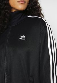 Svart zip-up jacka med hög krage, med tre vita ränder på varje axel och en Adidas-logotyp på den övre vänstra sidan.