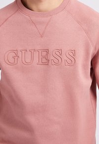 Nærbillede af en person iført en pink sweatshirt med hævet, tekstureret "GUESS"-bogstaver hen over brystet.