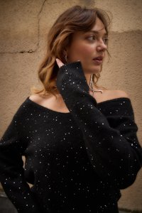 Juodas nuo peties megztinis su sidabrinėmis žvilgsnėmis, struktūruotas mezgimas, ilgomis rankovėmis ir ribotais rankogaliais prieš tamsų foną.