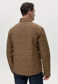 Giacca trapuntata marrone con colletto alto, caratterizzata da un tessuto testurizzato e dettagli di cucitura quadrata, abbinata a pantaloni scuri.
