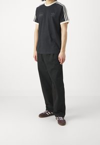 Osoba oblečená v černém tričku Adidas s bílými pruhy, volných černých kalhotách a hnědých teniskách Adidas stojící na bílé podlaze.