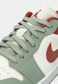 Gros plan sur les lacets et la languette de la sneaker présentant des superpositions gris-vert, un swoosh Nike rouge et un logo Jumpman rouge sur un fond en cuir blanc.