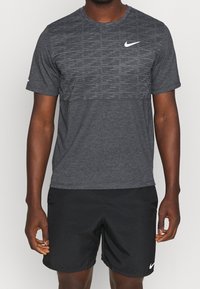 Szary sportowy t-shirt z teksturowaną górną częścią, krótkimi rękawami, okrągłym dekoltem i białym logo Nike. Noszony z czarnymi szortami.