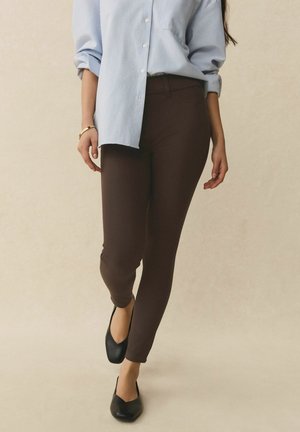 Frau geht und trägt schwarze spitze Ballerinas, braune eng anliegende Hose und ein blau gestreiftes, langärmeliges Hemd mit hochgekrempelten Ärmeln.