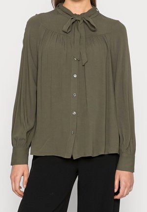 Femme portant un chemisier vert olive à manches longues avec des boutons sur le devant et un col noué en nœud, associé à un pantalon noir.