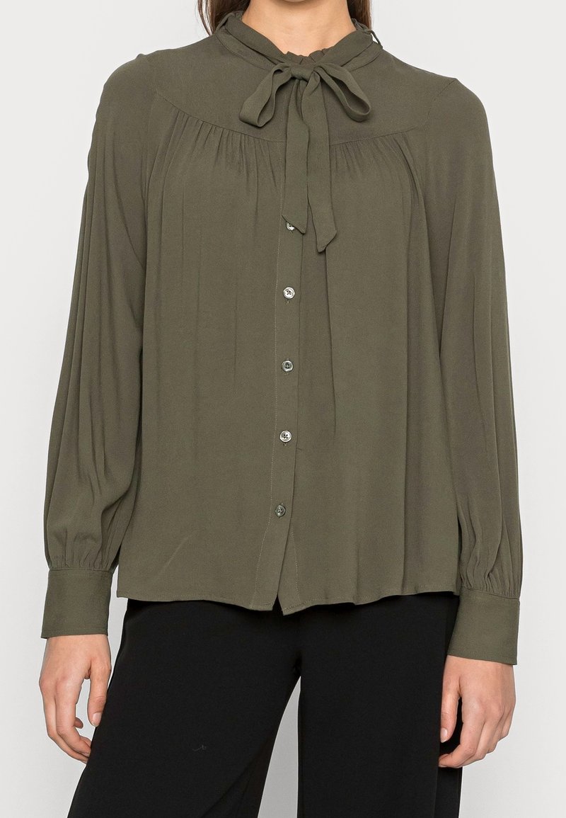 Femme portant un chemisier vert olive à manches longues avec des boutons sur le devant et un col noué en nœud, associé à un pantalon noir.