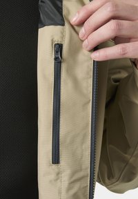 Hand, die eine beige Jacke hält, auf der Vorderseite sind zwei schwarze vertikale Reißverschlüsse zu sehen, darunter schwarzer, strukturierter Stoff.