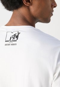 Camiseta de algodón blanca con un logo negro de MTV y el texto "MUSIC TELEVISION" arriba, con "ANTONY MORATO" abajo. Diseño clásico de cuello redondo.