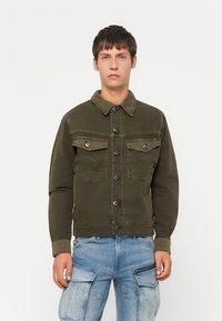 Jonge man met kort donker haar die een donkergroene, gebuttonte jas en lichtblauwe cargojeans draagt, staande tegen een effen achtergrond.