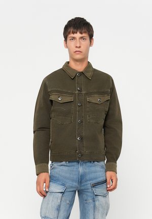 Jonge man met kort donker haar die een donkergroene, gebuttonte jas en lichtblauwe cargojeans draagt, staande tegen een effen achtergrond.