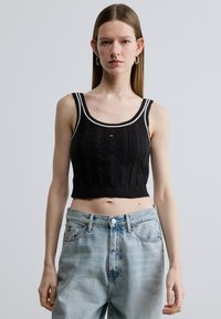 Juodas megztas trumpas viršus su tekstūruotu raštu, kuriame yra baltos akcentinės petnešėlės ir mažas logotipas priekyje. Dėvimas su šviesiai mėlynais džinsais.