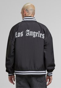 Svart bomberjacka med vit text "Los Angeles" på baksidan, randiga muddar och nederkant, ribbad krage, tillverkad av slät material.
