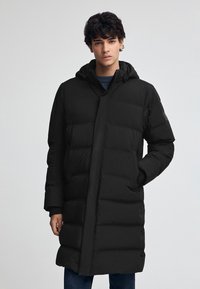 Svart puffjacka med huva, vertikal quiltad design, framfickor och dragkedjestängning. Materialet verkar glatt och isolerat.