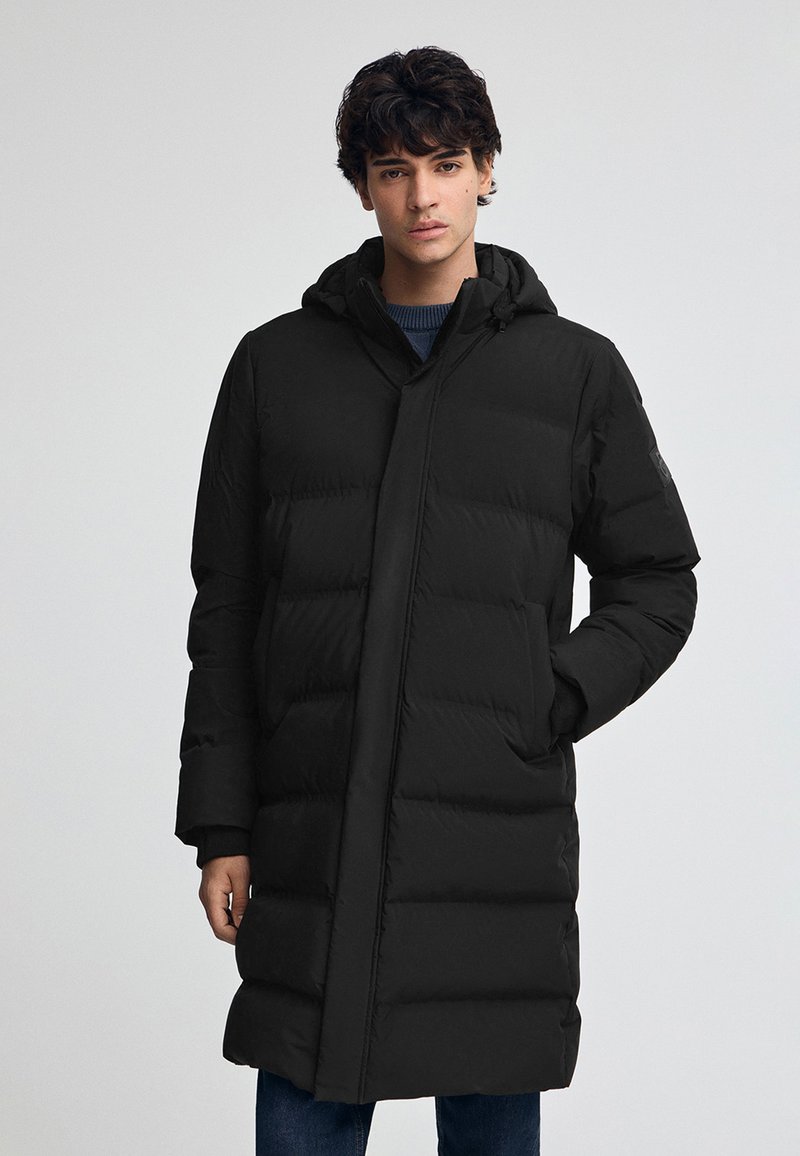 Svart puffjacka med huva, vertikal quiltad design, framfickor och dragkedjestängning. Materialet verkar glatt och isolerat.