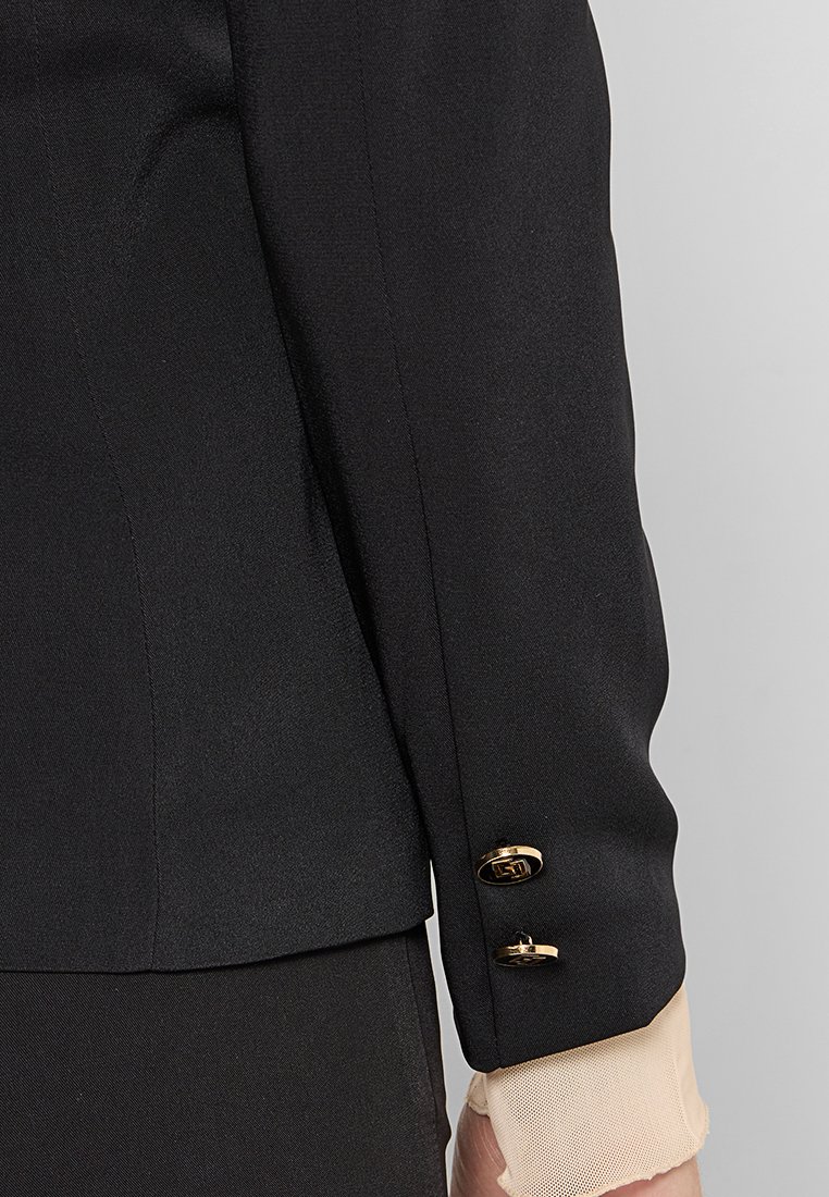Blazer negro con acentos de botones dorados, ajuste entallado y una textura suave, con un puño claro y transparente visible debajo.