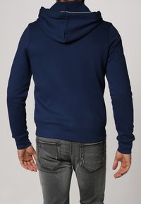 Marinblå hoodie i mjukt tyg, med dragsko i huvan och ribbat nederkant. Kombinerat med åtsittande grå jeans med diskreta detaljer på bakfickan.