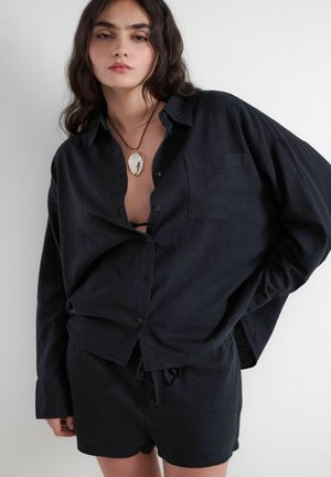 Woman with dark wavy hair wears loose black button-up shirt, matching shorts, and shell pendant necklace, standing against a white background.