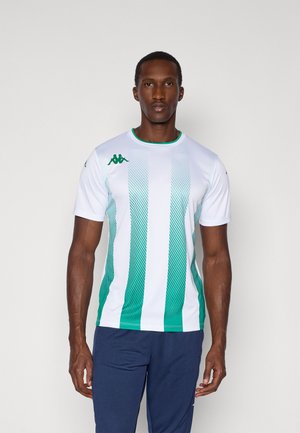 Uomo in piedi su sfondo bianco, indossa una maglia sportiva a righe bianche e verdi e pantaloni blu navy, volto rivolto in avanti con espressione neutra.