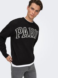 Black knit sweater featuring white "PARIS" lettering across the chest, ribbed cuffs, and hem. Worn over a white T-shirt and paired with gray jeans.