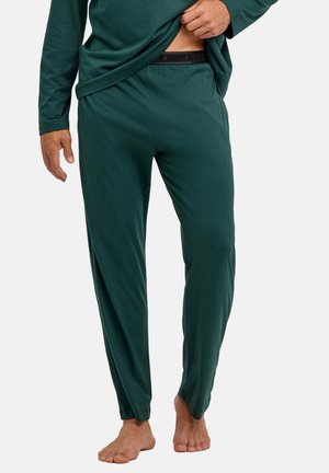 Man draagt een donkergroen shirt met lange mouwen en bijpassende broek, tilt het shirt iets op om de zwarte tailleband te tonen, staat blootsvoets op een witte achtergrond.