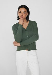 Cardigan verde con relieve, de manga larga y escote en V, que presenta dos botones en la parte delantera. Combinado con pantalones blancos.
