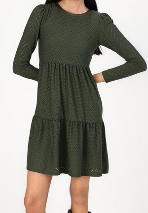 Kvinde iført en mørkegrøn, langærmet, mønstret knælængde kjole med en lagdelt nederdel og tætsiddende talje.