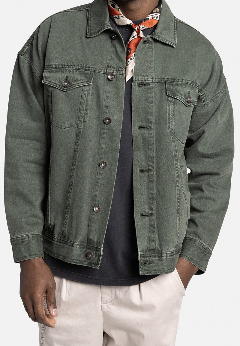 Groene spijkerjasje met een comfortabele pasvorm, twee borstzakken, knoopsluitingen en een gestructureerde stof. Draagt over een zwart T-shirt en gecombineerd met een sjaal.