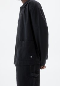 Black work jacket made of cotton, featuring a button-up design, large front pocket, and a small logo tag on the pocket.