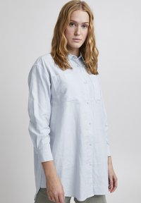 Chemise bleu clair à manches longues, en tissu texturé, dotée d'une poche poitrine et d'une coupe décontractée, rentrée dans un pantalon de couleur claire.