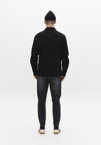 En man som bär en svart beanie, en svart långärmad skjorta, mörka jeans och vita sneakers, står vänd mot en vit bakgrund.