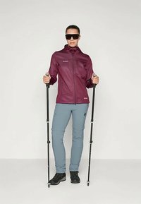 Maroon zip-up jacket, light grey pants, black hiking boots; holding trekking poles, wearing dark sunglasses, against a plain background.