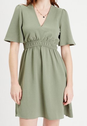 Femme portant une robe vert clair à manches courtes avec un décolleté en V et une ceinture élastique, accessoirisée d'un collier chaîne.