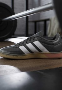 Zwart-witte sneaker op de gymvloer met een halter en gewichtsschijven in de vage achtergrond, onder zacht natuurlijk licht.