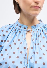 Γυναίκα που φοράει μπλε μπλούζα με καφέ πουά, δεμένο ντεκολτέ, κοντά καφέ μαλλιά και μικρό ορθογώνιο ασημένιο σκουλαρίκι στο αριστερό αυτί.