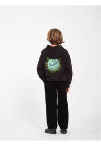 Zwarte hoodie met achterkant bedrukking, featuring groene tekst en zonnestraalontwerp, gecombineerd met zwarte corduroy broeken en zwarte schoenen, gezien van achteren.